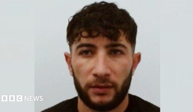 A police mugshot of a man with short black hair and a black beard who is wearing a black t-shirt