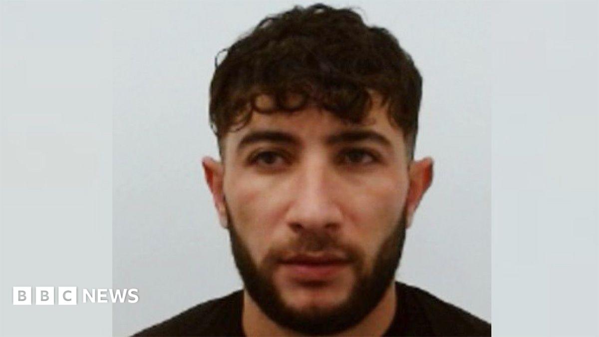 A police mugshot of a man with short black hair and a black beard who is wearing a black t-shirt