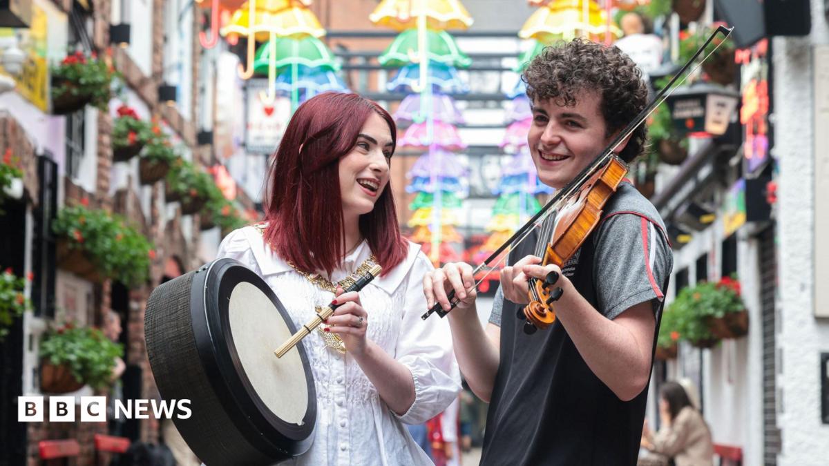 A woman and man standing in Belfast's Cathedral Quarter. The woman, with dark red hair, is holding a bodhrán. The man, with short, curly brown hair, is holding a fiddle.