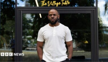 A young black man wearing a white T-shirt and standing outside in front of glass doors