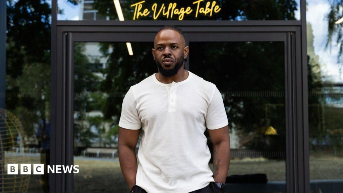 A young black man wearing a white T-shirt and standing outside in front of glass doors