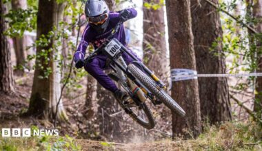 A person is mid-air as they ride their bike through the forest. They are wearing a purple outfit, plus a grey helmet and dark sunglasses. There are tall trees in the background. One on the right has white tape wrapped around it.