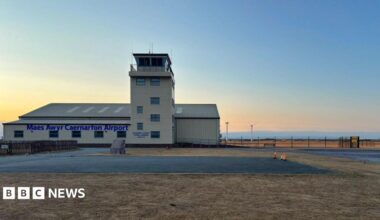 A small cream building airport. It has a watchtower and surrounded by green grass and fields. It also has car parks all around.