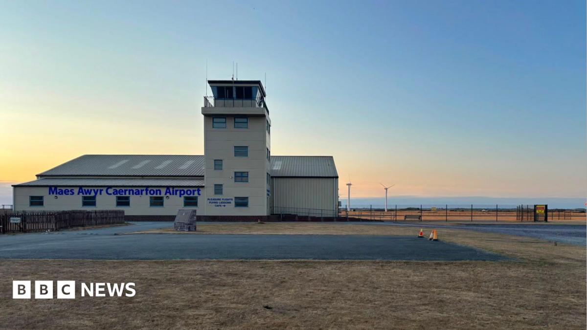 A small cream building airport. It has a watchtower and surrounded by green grass and fields. It also has car parks all around.