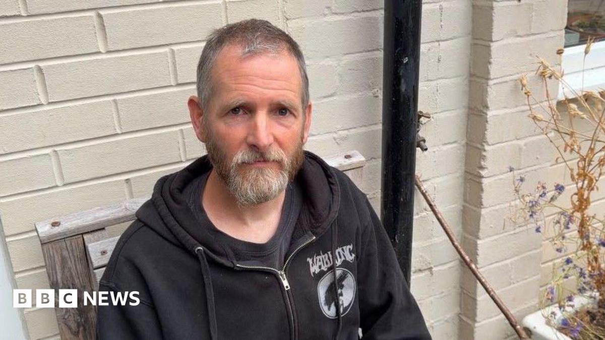A man with a grey beard and short hair wearing a black hooded top and sitting outside on a wooden chair.