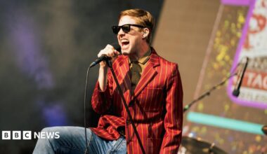 Ricky Wilson of the Kaiser Chiefs, dressed in jeans and a yellow striped red blazer, wearing a yellow shirt and sunglasses, singing into a leaning microphone stand.