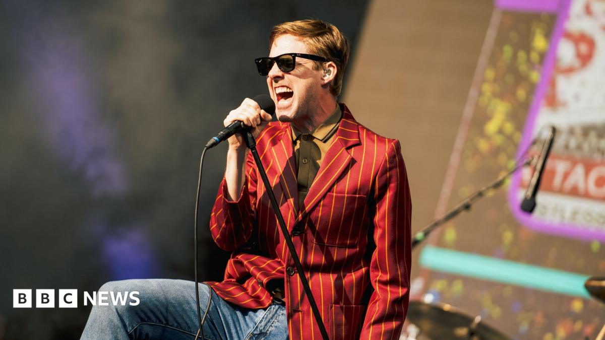 Ricky Wilson of the Kaiser Chiefs, dressed in jeans and a yellow striped red blazer, wearing a yellow shirt and sunglasses, singing into a leaning microphone stand.