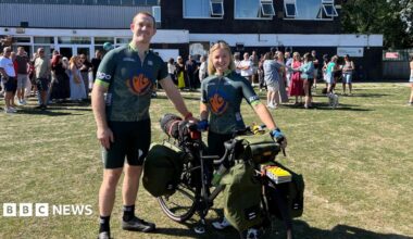 Lucy and Ciaran stand together with a bike that is loaded with panniers, wearing matching, green cycling jerseys. It is a sunny day and they are standing on grass, with a crowd of people mingling in the background.