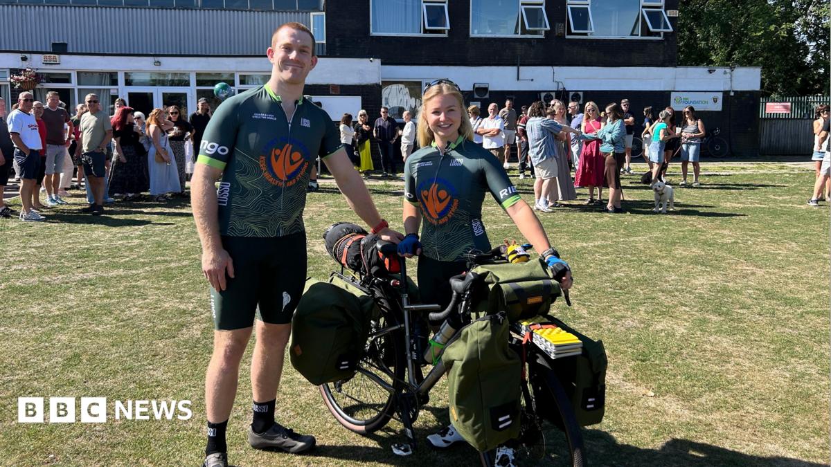 Lucy and Ciaran stand together with a bike that is loaded with panniers, wearing matching, green cycling jerseys. It is a sunny day and they are standing on grass, with a crowd of people mingling in the background.