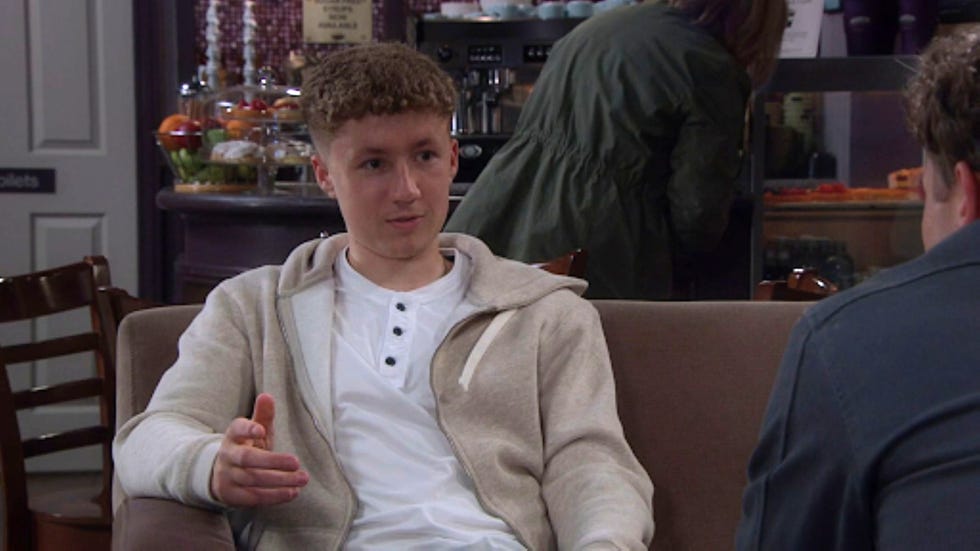 emmerdale noah dingle person in casual attire sitting on a sofa, gesturing with one hand in a café setting.