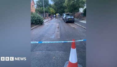 A traffic cone and police tape can be seen at one side of a road, with some water seen on the road and an emergency services vehicle at the other end of the road.