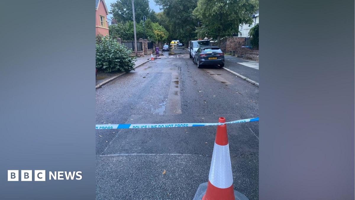 A traffic cone and police tape can be seen at one side of a road, with some water seen on the road and an emergency services vehicle at the other end of the road.