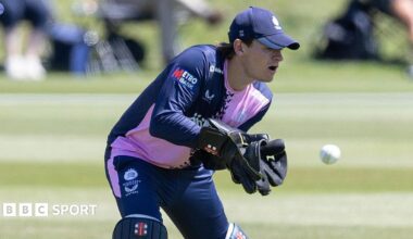 Jack Davies keeping wicket in gloves, cap and pads, with navy and pink shirt and navy trousers