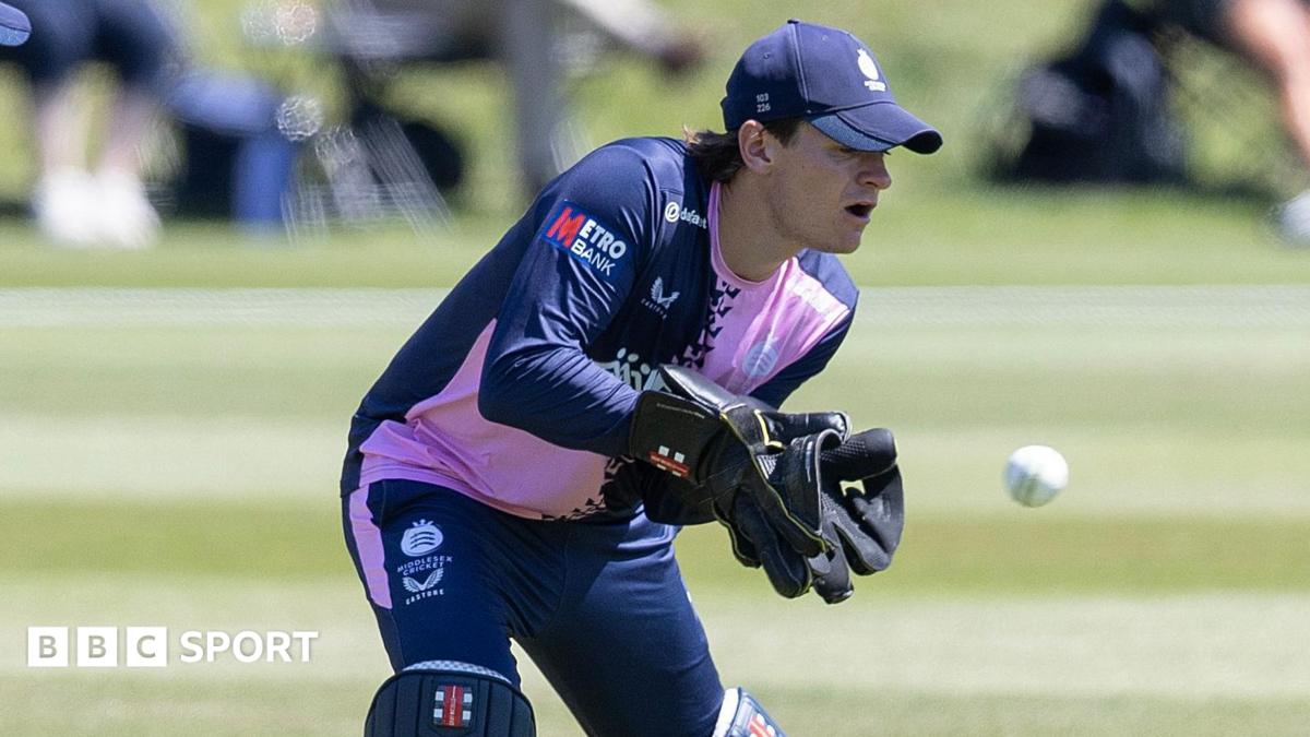 Jack Davies keeping wicket in gloves, cap and pads, with navy and pink shirt and navy trousers