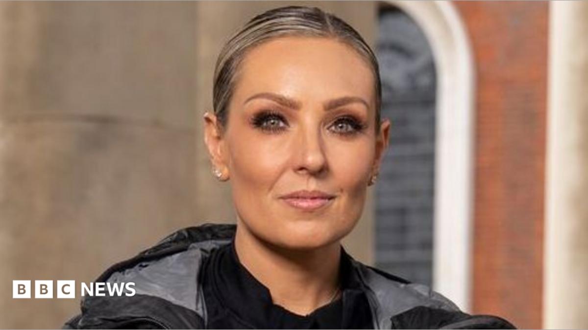 Carlos Gu and Amy Dowden pictured side by side in a promotional photo for the Channel 4 show, Celebrity Hunted. Carlos is wearing a grey dark coat, a light grey t-shirt and several necklaces. Amy is wearing a black top, jacket and coat. They are both looking at the camera and are not smiling.