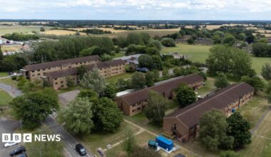Aerial shot of the housing blocks at an airbase. Four rectangular accommodation blocks can be seen, surrounded by grass, trees and roads. There are agricultural fields in the background.
