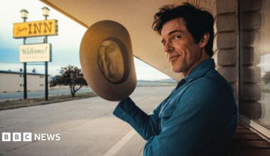 Brandon Flowers holds a stetson in his right hand and gives a wry look to the camera as he poses outside a stereotypical American motel