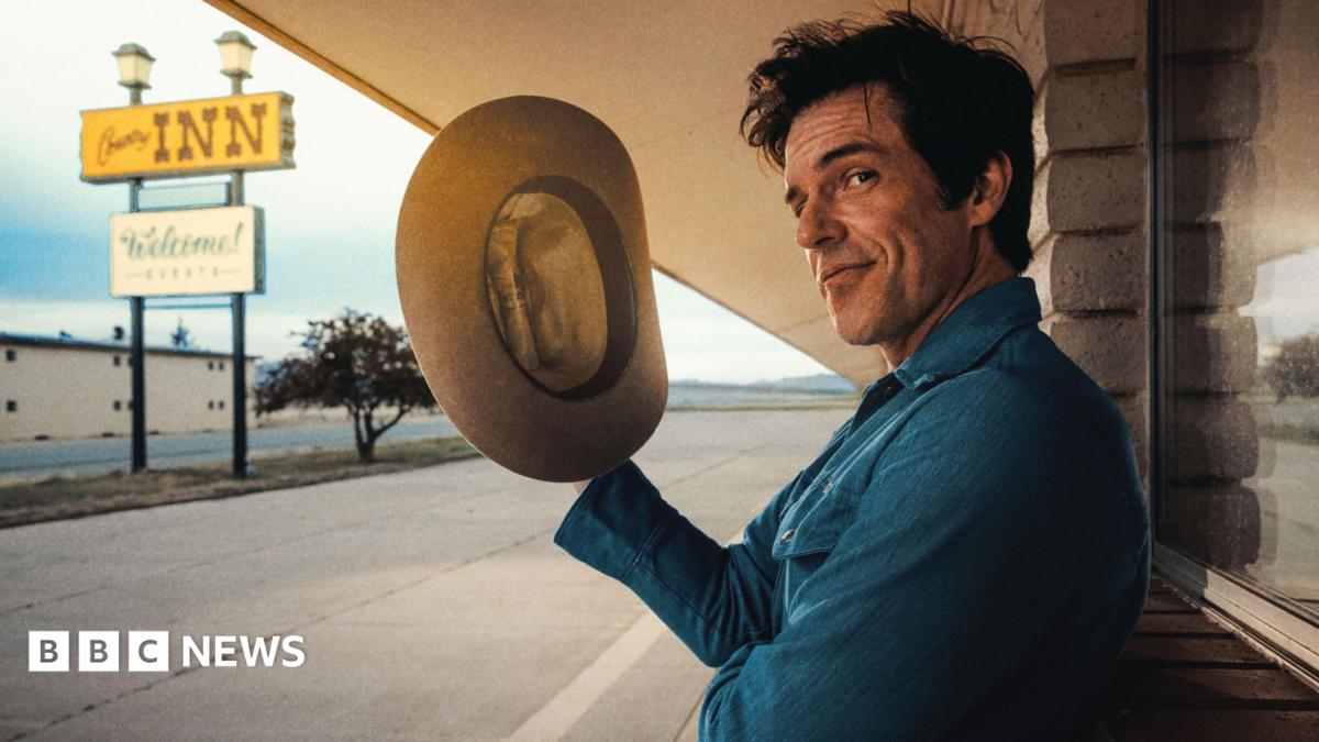 Brandon Flowers holds a stetson in his right hand and gives a wry look to the camera as he poses outside a stereotypical American motel