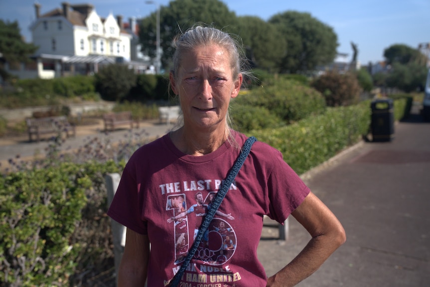 A 60 year old woman with a burgundy t shirt and grey hair in a pony tail stands on a residential street.