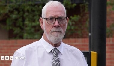 A close-up  of a Gerry Duffy seen  with short white hair, a neat white beard and moustache, and black-rimmed glasses looking directly at the camera with a somber expression. He is wearing a formal white collared shirt and a diagonal grey and white striped tie. The background shows a blurred outdoor setting with a red brick wall, green foliage, and a dark pole.