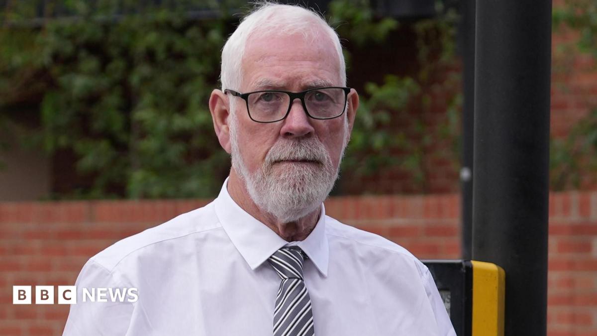 A close-up  of a Gerry Duffy seen  with short white hair, a neat white beard and moustache, and black-rimmed glasses looking directly at the camera with a somber expression. He is wearing a formal white collared shirt and a diagonal grey and white striped tie. The background shows a blurred outdoor setting with a red brick wall, green foliage, and a dark pole.