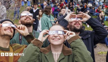 A group of people all holding special glasses to their faces and looking up at the sky to view a solar eclipse. They are wearing coats and in the background are crowds of people standing or sitting on a patch of grass.