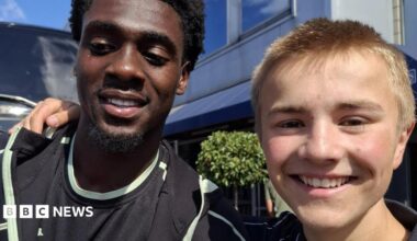 Norwich striker Mo Touré (left) is wearing a black T-shirt. He is standing next to Ranulph Bacon, who is wearing a navy polo shirt. The pair are smiling at the camera.