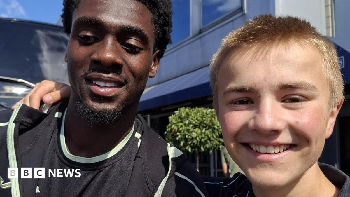 Norwich striker Mo Touré (left) is wearing a black T-shirt. He is standing next to Ranulph Bacon, who is wearing a navy polo shirt. The pair are smiling at the camera.