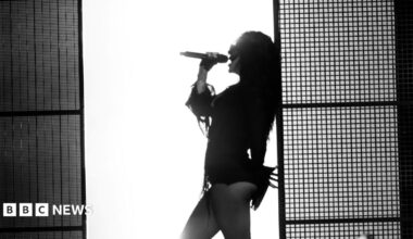 A black and white photo of Charli XCX leaning against a wall as she sings into a microphone. There is a bright white wall behind her which silhouettes her body.