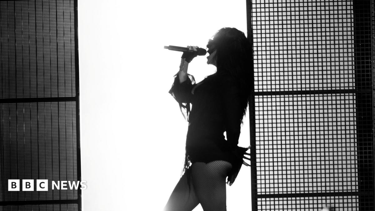 A black and white photo of Charli XCX leaning against a wall as she sings into a microphone. There is a bright white wall behind her which silhouettes her body.
