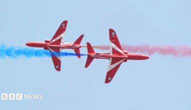 Two Red Arrow planes move away from each other in the sky, one releasing blue smoke and the other red smoke.