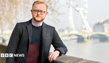 A man in a black jacket and with glasses, ginger hair and a beard stands on the riverside by the River Thames with Westminster Bridge and the London Eye in the background.