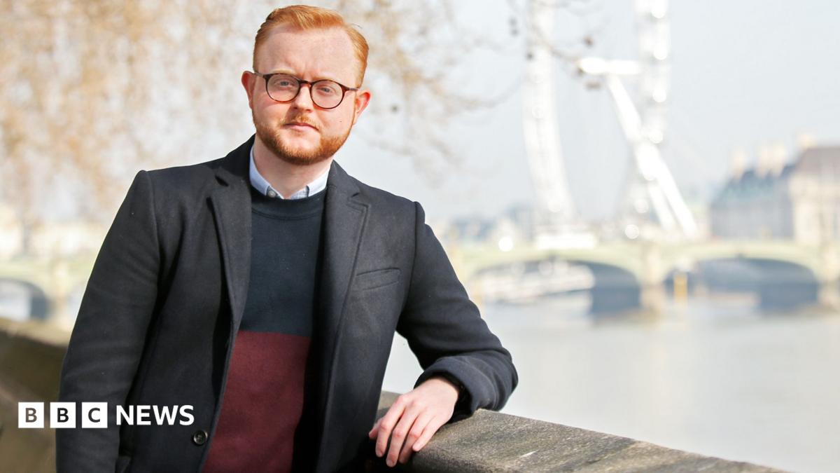 A man in a black jacket and with glasses, ginger hair and a beard stands on the riverside by the River Thames with Westminster Bridge and the London Eye in the background.