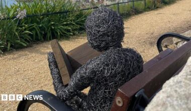 A metal sculpture of a human figure reading a book on a wooden park bench
