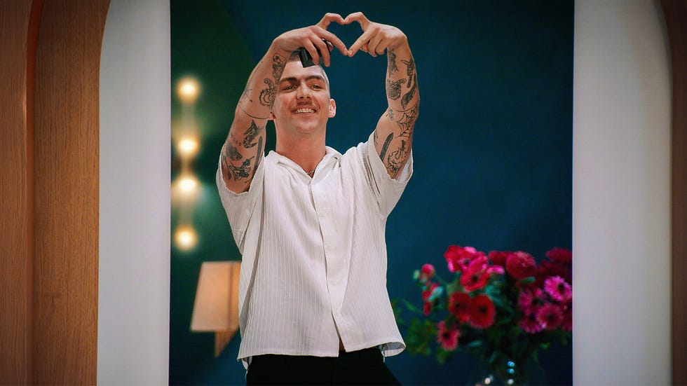 Love is blind uk jack and faye person making a heart shape with hands, surrounded by tattoos, flowers in background.