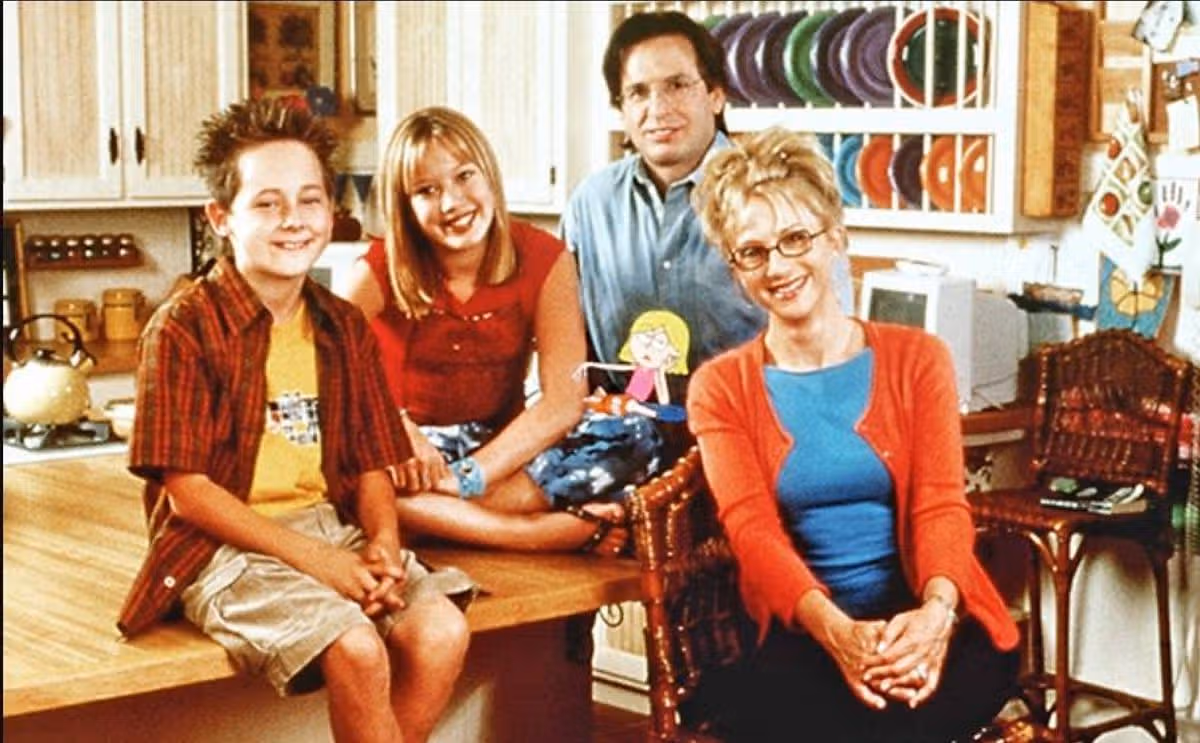 An image of a family of four individuals seated together in a kitchen, with a wooden table and shelves in the background.