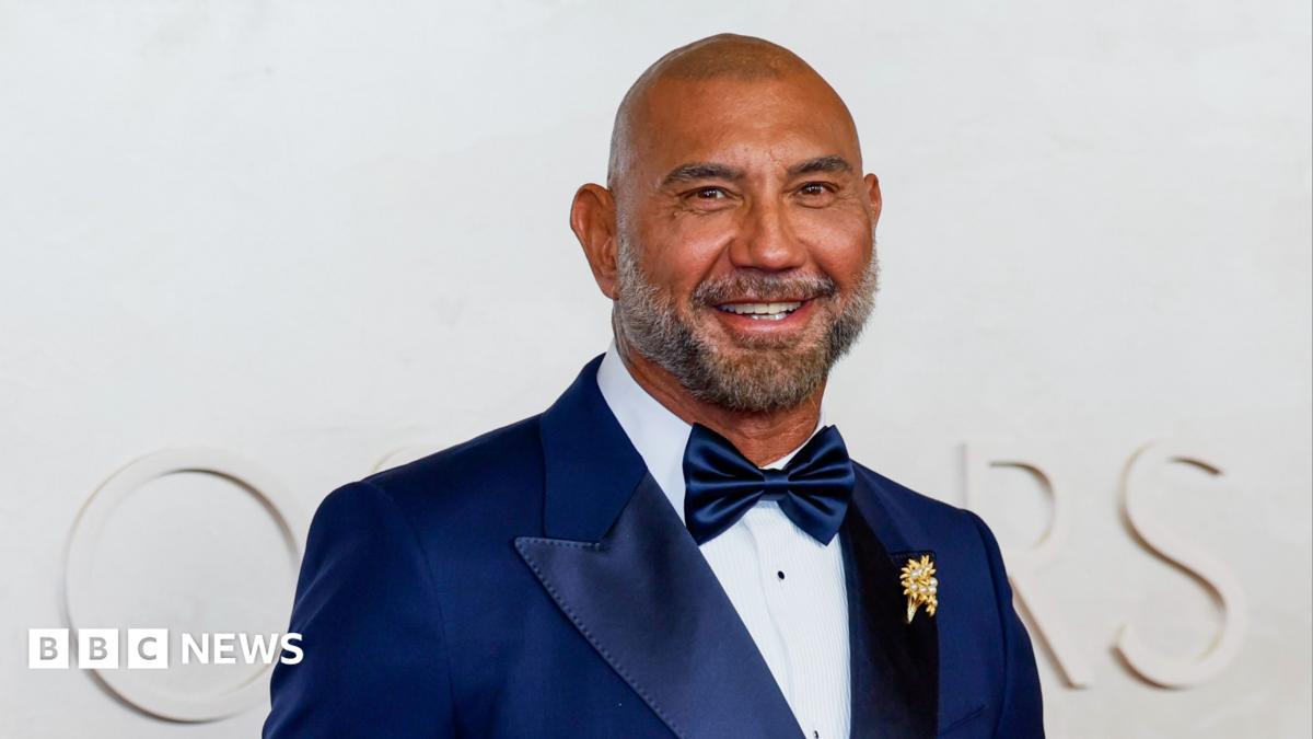 Dave Bautista smiling and looking at the camera on a red carpet. He's wearing a tux with a white shirt, blue bow tie and blue jacket. He's a bald man with a darker beard that's going grey down the sides.