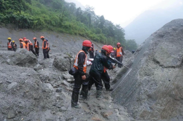 Workers in high vis and helmets use a large drill into a hard surface. They are in a valley where the ground is thick with mud and clouds hang in the air