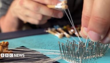 A close-up shot of hands working on bobbin lace, guiding fine white threads through rows of metal pins pinned into a turquoise cushion.