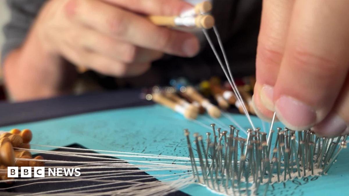 A close-up shot of hands working on bobbin lace, guiding fine white threads through rows of metal pins pinned into a turquoise cushion.