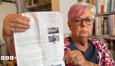 A woman with short blonde and pink hair and pink-rimmed glasses and a multi-coloured top is holding up a large piece of paper, which is a parking fine, at the camera. There is a bookshelf in the background.