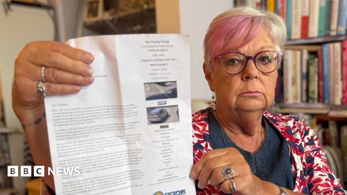 A woman with short blonde and pink hair and pink-rimmed glasses and a multi-coloured top is holding up a large piece of paper, which is a parking fine, at the camera. There is a bookshelf in the background.
