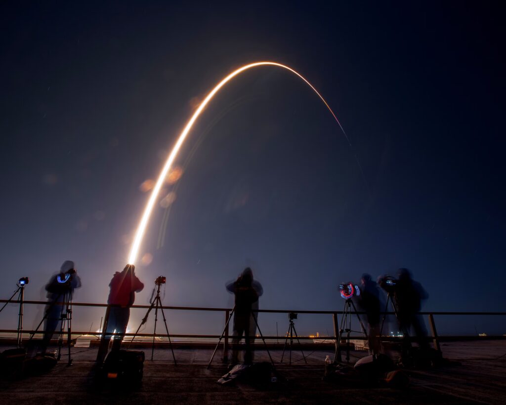 Photographers gathered on a balcony as a distant bright flash arcs through a dark sky. 