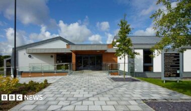 The Castlegate and Derwent Surgery site. It is a modern-looking building and the entrance has glass doors. A sign to the right-hand side of the doors directs visitors to different departments. A young tree planted next to the sign.