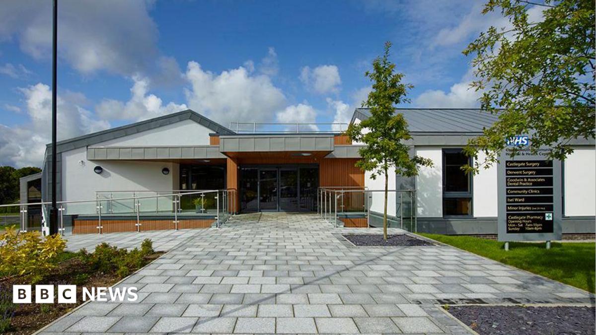 The Castlegate and Derwent Surgery site. It is a modern-looking building and the entrance has glass doors. A sign to the right-hand side of the doors directs visitors to different departments. A young tree planted next to the sign.