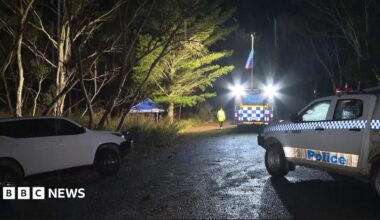 A night scene on a country road with several police cars and a small blue tent roof behind some trees.