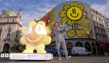 Mr Doodle stands beside one of his sculptures in Piccadilly Circus. The installation features his signature black-and-white doodle artwork with bright orange and yellow colours, while an image of the artist appears on the famous Piccadilly Circus screen behind him