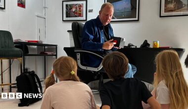 The image shows a man sat on a chair talking to a group of five children who are all sat on the floor. Framed pictures can be seen on the wall in the background.