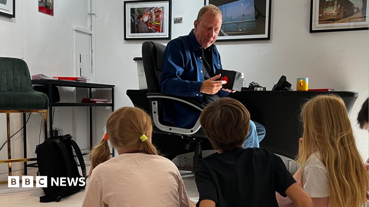 The image shows a man sat on a chair talking to a group of five children who are all sat on the floor. Framed pictures can be seen on the wall in the background.