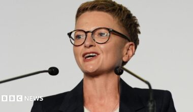Bev Craig has short spiky red hair, dark glasses and wears a dark jacket with white roll-neck top.